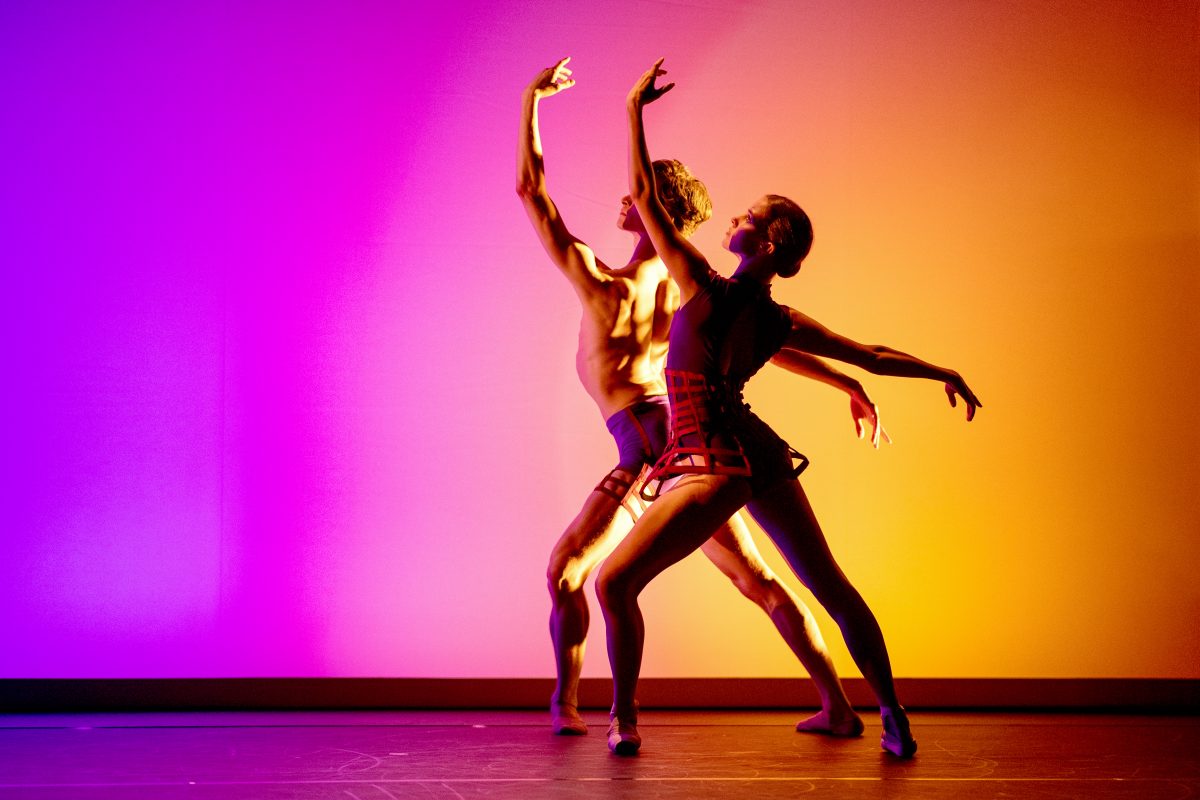 Scene from a pas-de-deux on stage, against purple and yellow lighting.